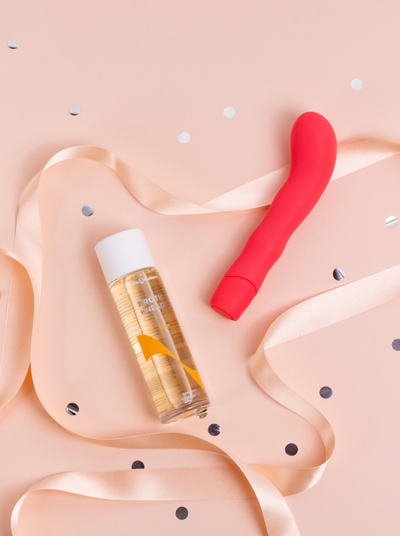 A red vibrator and massage oil on a pink background with ribbon and confetti